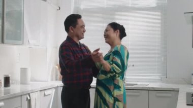 Yetişkin bir Asyalı çiftin ev mutfağında dans edip sohbet ederken birbirlerine baktıkları orta boy bir fotoğraf.