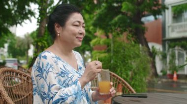 Orta yaşlı, Asyalı bir kadının açık hava kafesinde portakal suyu içtiği bir fotoğraf.