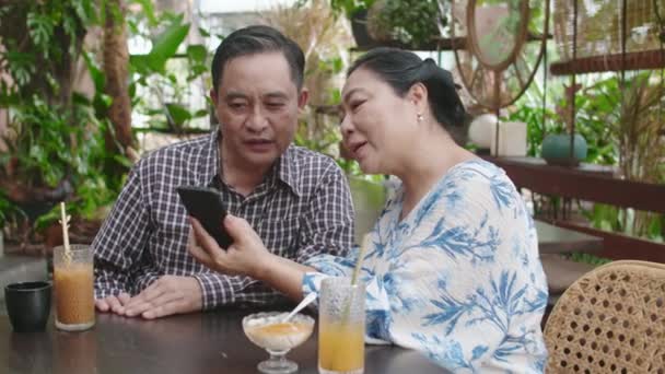 夫にカフェで飲み物を持ってテーブルに座って携帯電話を使用するように教えるアジアの女性のミディアムショット — ストック動画