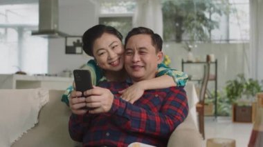 Evde akıllı telefondan selfie çekmeyi öğrenirken, Asyalı mutlu, yaşlı eşlerin birbirlerine sarıldıkları orta boy bir fotoğraf.