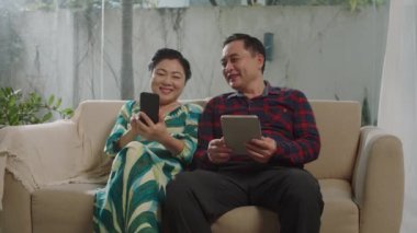 Orta yaşlı Asyalı karı ve kocanın akıllı telefon ve dijital tablet kullanarak evdeki kanepede otururken orta uzunlukta bir fotoğrafı.
