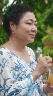 Asya kökenli yaşlı bir kadının dikey portresi açık kafede meyve suyu içiyor.