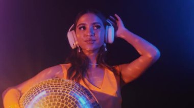 Neşeli Asyalı bir kadının disko topu tutarken, neon ışıkta kulaklıkla müzik dinlerken ve kameraya bakarken orta boy bir fotoğrafı.