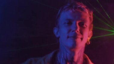 Boyunda dövmesi ve küpesi olan neon ışıklı bir gece kulübünde eğlenen neşeli genç bir adam.