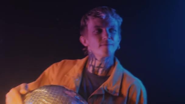 ネオンライトの屋内でディスコボールクラブを保持する首にタトゥーを持つ若い男性Djの中型ショット — ストック動画