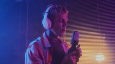 Neon Stüdyoda mikrofonda şarkı söyleyen genç beyaz erkek müzisyenin orta boy çekimi.