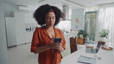 Turuncu gömlekli, akıllı telefon kullanan ve evden kameraya bakan neşeli kadın girişimcinin portresi.
