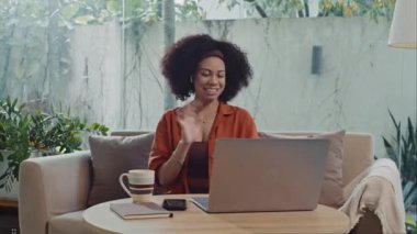Latin iş kadınının online toplantı sırasında iş arkadaşlarını karşılaması evdeki kanepede dizüstü bilgisayarın önünde oturması.