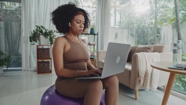 在家里 穿着运动服坐在健身球上的女自由职业者在笔记本电脑上工作的中长镜头 — 图库视频影像
