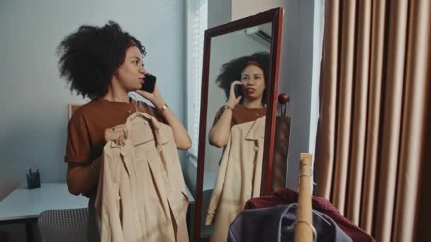 自宅の鏡の近くでベージュコートを試している間 携帯電話で話している若いカーリー女性の中長いショット — ストック動画