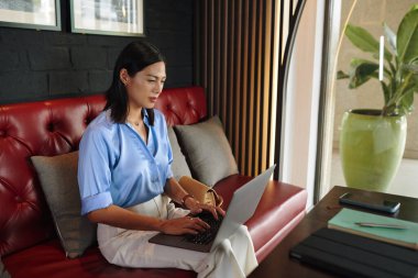 Kafede kanepede oturan ve dizüstü bilgisayarla çalışan bir kadın.