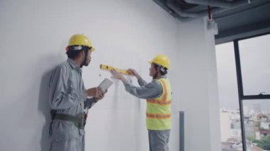 Orta uzunluktaki Asyalı ustabaşının duvarları ölçerken Afrikalı Amerikalı inşaatçıyla inşaat alanında çalışan bir aletle orta uzunlukta görüntüsü.