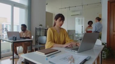 Genç Asyalı grafik tasarımcının diğer iş arkadaşlarıyla birlikte laptopta çalışırken geniş açılı görüntüsü