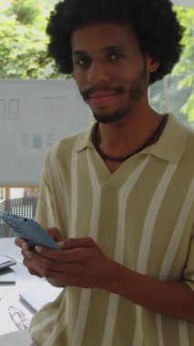 Akıllı telefonu tutarken kameraya gülümseyen Afro-Amerikan ofis çalışanının dikey portresi