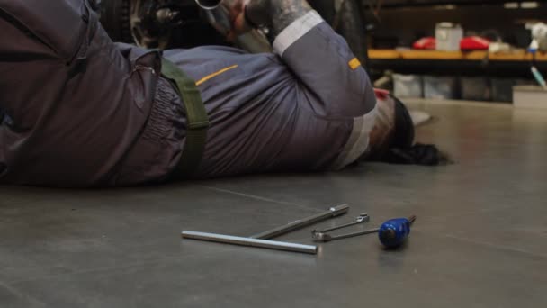 摩托车修理时躺在地板上 身穿灰色制服更换器械的多文化技师 — 图库视频影像
