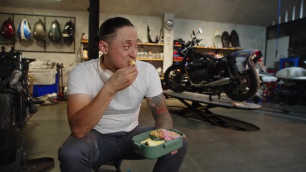Średnie Ujęcie Wielokulturowego Rowerzysty Jedzącego Lunch Plastikowego Pojemnika Garażu Motocyklowym — Wideo stockowe