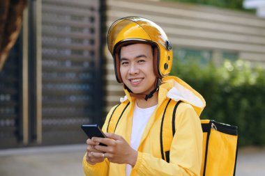 Sarı üniformalı gülümseyen Vietnamlı teslimatçının portresi mobil uygulama üzerinden müşterilerin siparişlerini kontrol ediyor.