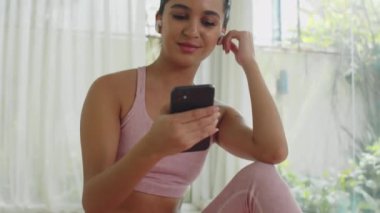 Kulaklıklı neşeli Asyalı kadının evde antreman yaptıktan sonra cep telefonundan komik videolar izlediği görüntüler.
