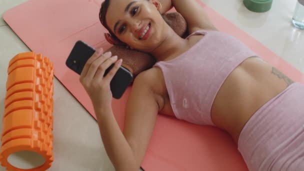 年轻的亚洲女孩在室内锻炼时躺在垫子上用手机嘲笑迷因的手持照片 — 图库视频影像