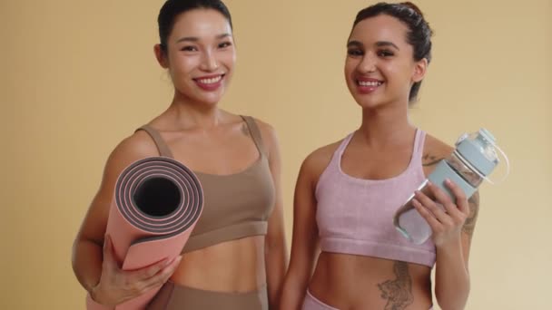 两名亚洲女运动员举着瑜伽垫和瓶装水 同时在黄色背景下摆好姿势拍照时的倒立肖像画 — 图库视频影像