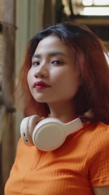 Açık havada poz verirken kulaklık takan turuncu tişörtlü Asyalı kızın dikey portresi.