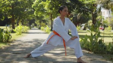 Tekvando dersinde açık havada antrenman yaparken beyaz takım elbiseli, turuncu kuşaklı genç Asyalı bir kızın uzun pozu.