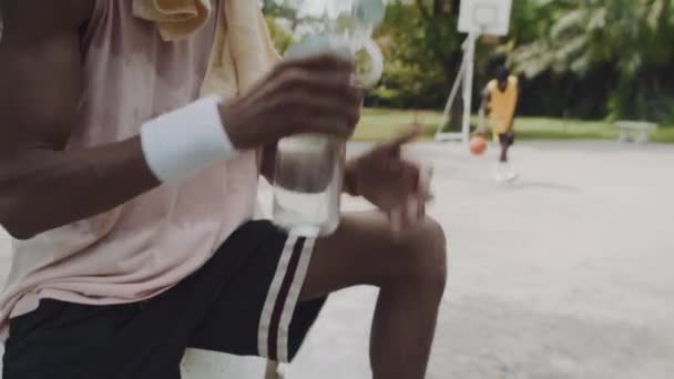 アウトドアトレーニング後に休憩しながら飲んでいる男性のブラックバスケットボール選手のチルトアップショット ロイヤリティフリーストック映像