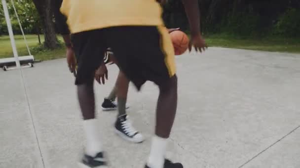 屋外の遊び場でのストリートボールトレーニング中に彼の対戦相手バウンスボールをオフフフロアで攻撃するブラックスポーツマンのハンドヘルドショット ストック映像