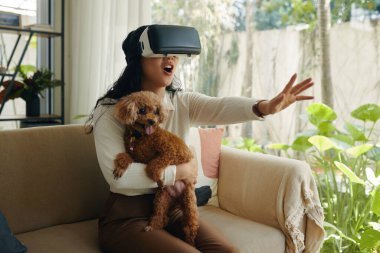 Video oyunu oynarken VR gözlükleriyle köpeğini tutan heyecanlı genç kadın