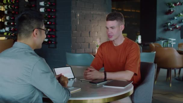 レストランのデジタルタブレットでオープンな財務チャートでテーブルに座って取引をする2人の多様なトレーダーの中型ショット — ストック動画