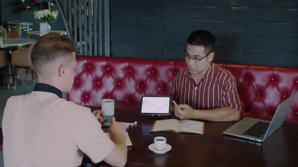 カフェのテーブルでデジタルタブレットの画面を指す株式について男性アジアの専門家に尋ねる若い白人トレーダーのミディアムショット — ストック動画