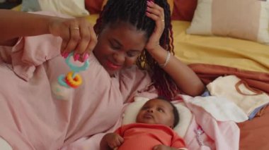 Siyah annenin bebek kafasında çıngırak sallayıp evde yatakta yan yana yattığı orta boy bir fotoğraf.