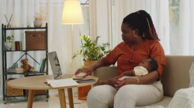 Dizüstü bilgisayarın önünde oturan, evde çalışırken uyuyan yeni doğmuş bir kızı tutan siyah kadının yan görüntüsü