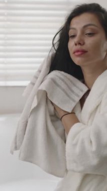 Bornozlu esmer bir kadının banyoda havluyla saçlarını silerken dikey pozu.