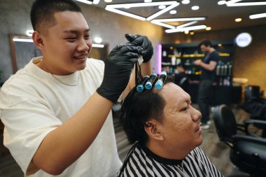 Gülümseyen berber müşterinin ıslak saçlarına bigudiler takıyor.