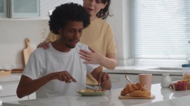 Orta boy beyaz bir kız, Afrika kökenli Amerikalı erkek arkadaşına sarılıp ev mutfağında kahvaltı ederken.