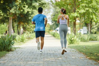 Erkek ve kadın güneşli bir günde parkta birlikte koşuyorlar, ikisi de spor kıyafeti giyiyorlar.