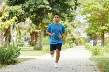 Parlak bir günde arka planda ağaçlarla çevrili yeşil bir yolda koşan bir adam.