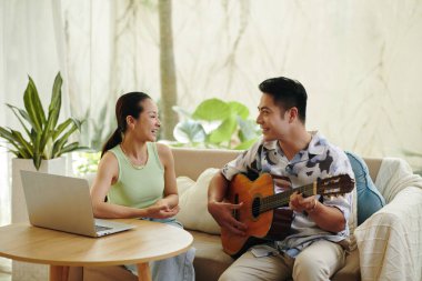 Evde gitar çalan bir adam ve gülümseyen bir kadınla, doğal ışıkla aydınlık bir oturma odasında güzel vakit geçiren bir çift.