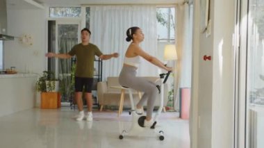 Uzak çekim Asyalı adam ip atlarken İspanyol kökenli kız arkadaşı evde antrenman yaparken spor bisikletine binerken.