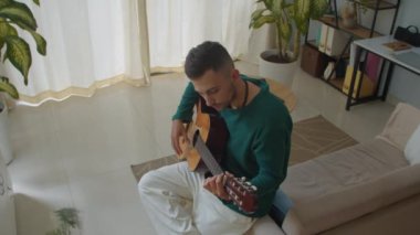 Kafkasyalı erkek söz yazarının evde akustik gitarla melodi yaratırken orta uzunlukta bir çekimi.