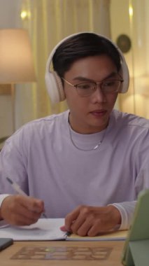 Ev ödevi yaparken kulaklık takan Asyalı bir gencin dikey fotoğrafı.