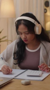 Kulaklıklı Asyalı bir kızın matematik ödevi yaparken, hesap makinesi kullanırken, evde masa başında otururken dikey bir fotoğrafı.