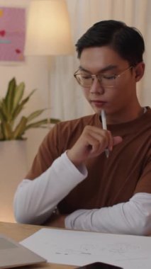 Asyalı adamın evden ders çalışırken proje kağıdına not aldığı dikey bir fotoğraf.