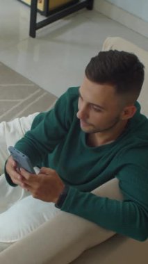 Evdeki kanepede otururken, beyaz erkek girişimcinin akıllı telefonuyla tele-sekreter çalışmasının dikey görüntüsü