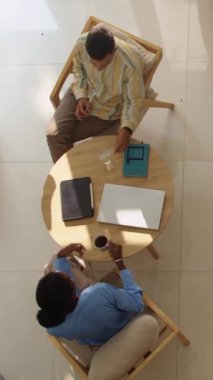 Modern ofisteki dinlenme yerinde kahve molası sırasında çift ırklı iş arkadaşlarının sohbetlerinin dikey yüksek açılı görüntüsü