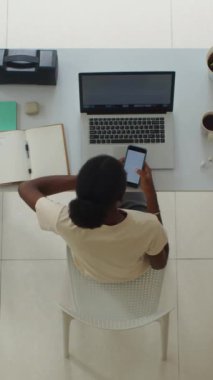 Siyahi iş kadınının ofis masasında otururken akıllı telefondan programı kontrol edişinin dikey üst görüntüsü