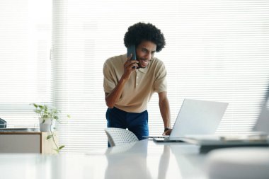 Gülümseyen işadamı telefona cevap verirken masaya yaslanıyor ve laptoptaki e-postaları kontrol ediyor.