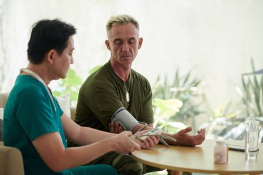 Doktor kan basıncını ölçmek için cihaz kullanırken asker masada oturuyor