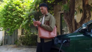Erkek Hintli girişimcinin EV 'nin yanında açık hava limanında kabloyla şarj olurken akıllı telefondan mesajlaşırken elde çekilmiş bir fotoğrafı.